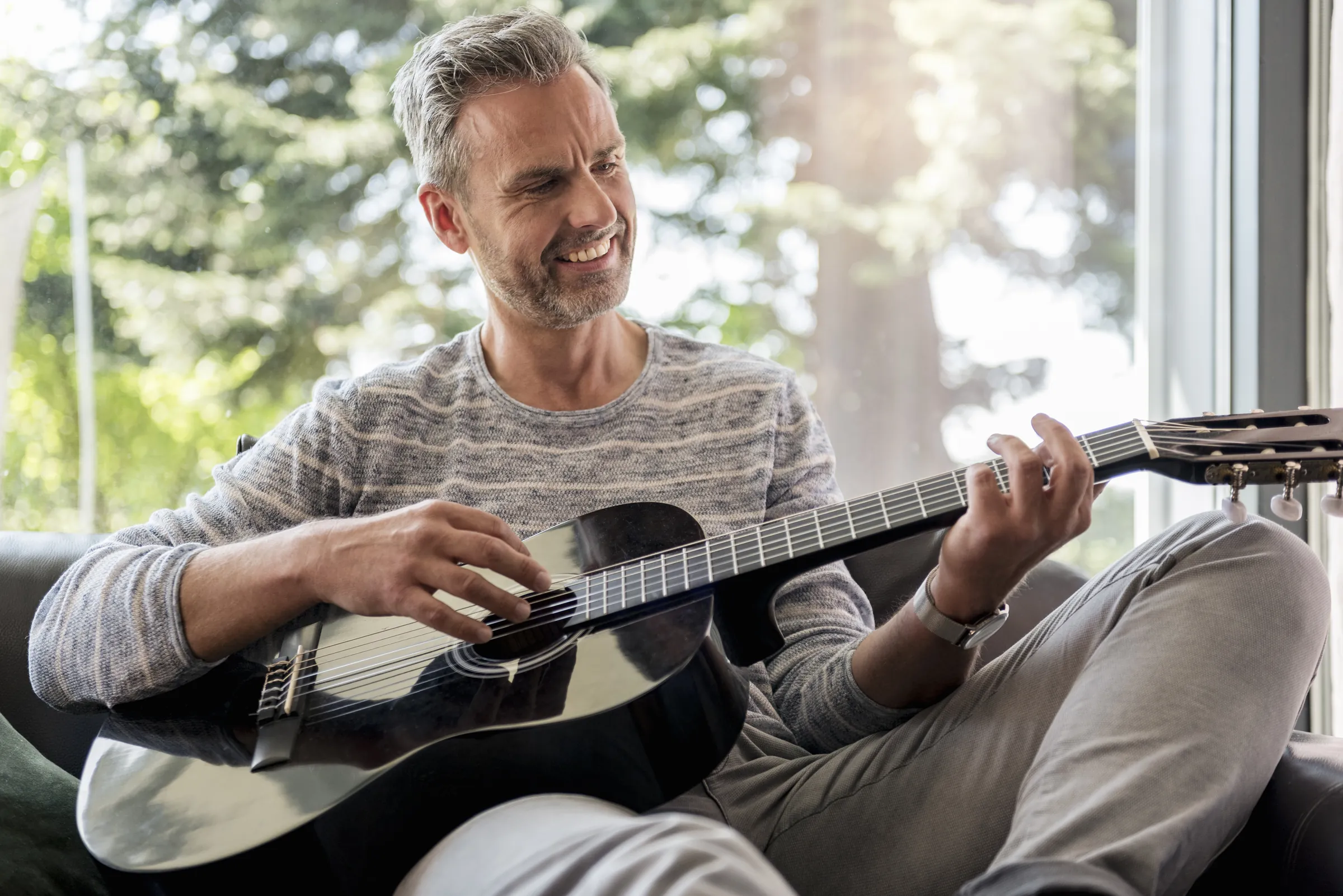 Hombre de mediana edad sonríe mientras toca la guitarra sentado en el sofá de su casa