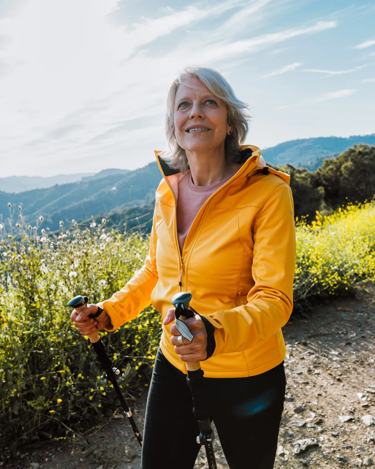 Mujer de mediana edad camina sonriente con bastones de senderismo por un camino de montaña