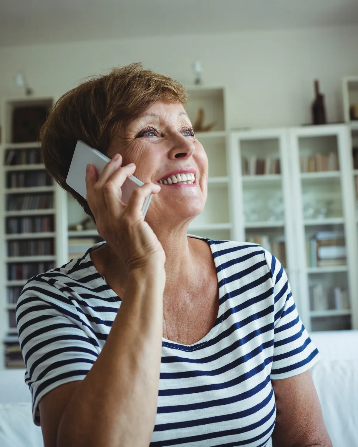Mujer de mediana edad sonríe mientras habla por teléfono sentada en el sofá de su casa