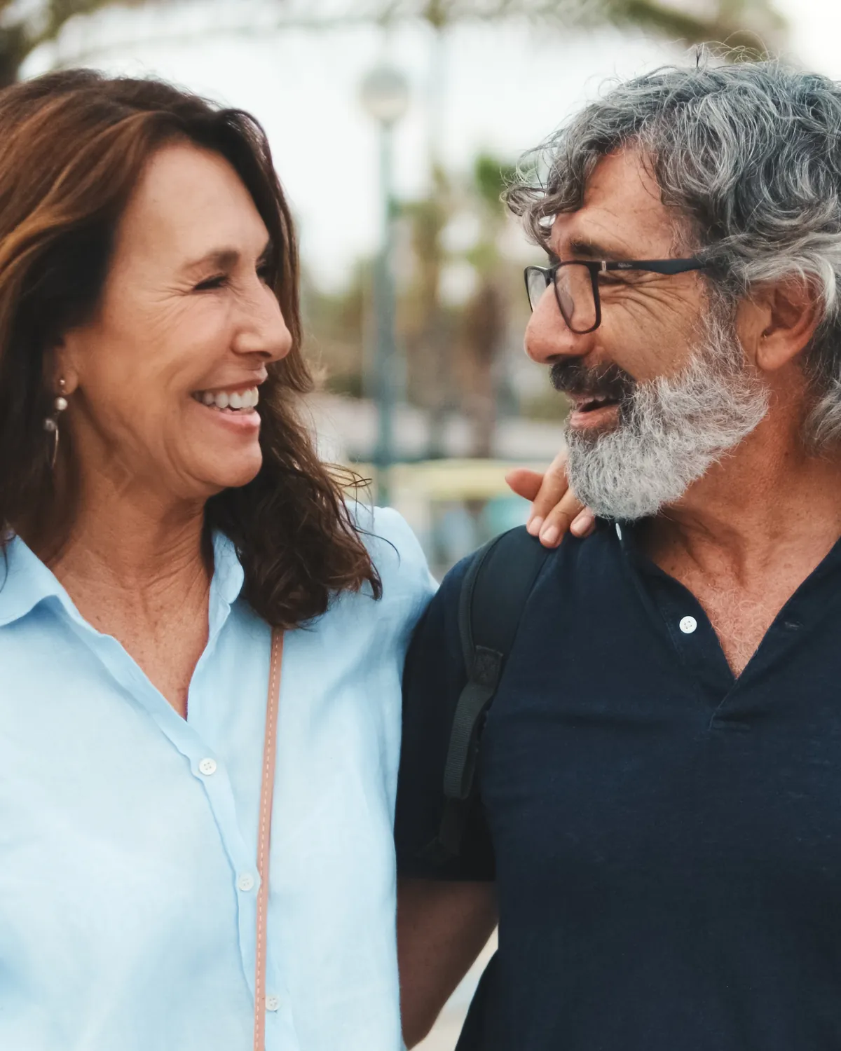 Una mujer y un hombre de mediana edad pasean abrazados por un paseo con palmeras y se miran sonriendo